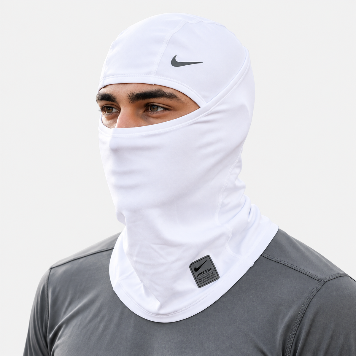 A man wearing a white Nike balaclava face covering with a dark gray Nike swoosh on the forehead and a small black Nike logo patch on the neck, paired with a dark gray athletic shirt against a light gray background.